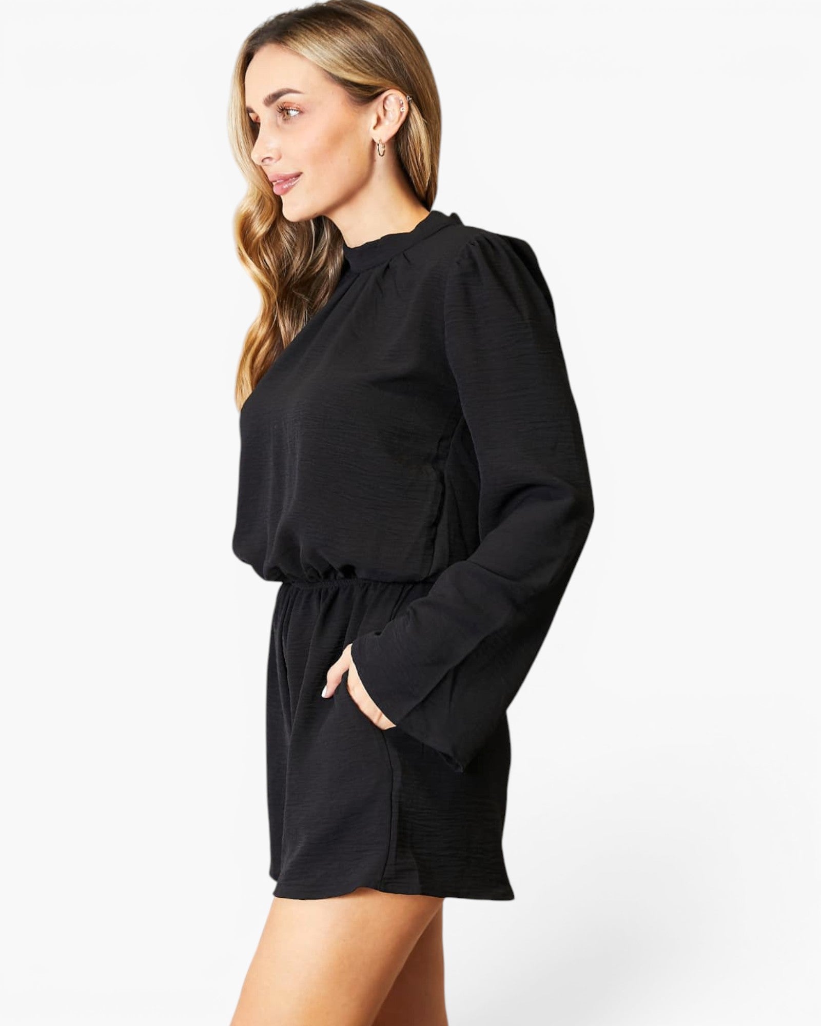 A woman wearing a black long-sleeve mini dress with a gathered neckline, puffed sleeves, and a drawstring waist, photographed against a clean white studio background.