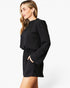 A woman wearing a black long-sleeve mini dress with a gathered neckline, puffed sleeves, and a drawstring waist, photographed against a clean white studio background.