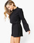 A woman modeling a black long-sleeve romper with a gathered high neckline and a matching fabric tie belt at the waist, photographed against a plain white studio background.