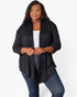 A woman modeling a black open-front cardigan with three-quarter sleeves, worn over a white tank top and blue jeans, against a plain white studio background.