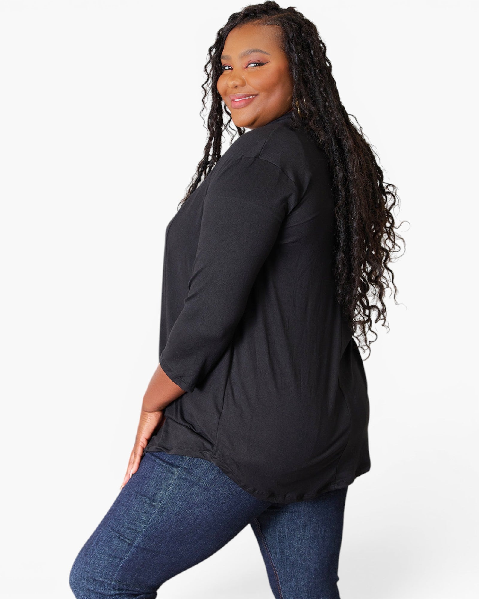 A woman with long, curly black hair is shown from a side-back angle wearing a loose-fitting black three-quarter sleeve top and blue jeans against a plain white studio background.