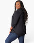 A woman with long, curly black hair is shown from a side-back angle wearing a loose-fitting black three-quarter sleeve top and blue jeans against a plain white studio background.