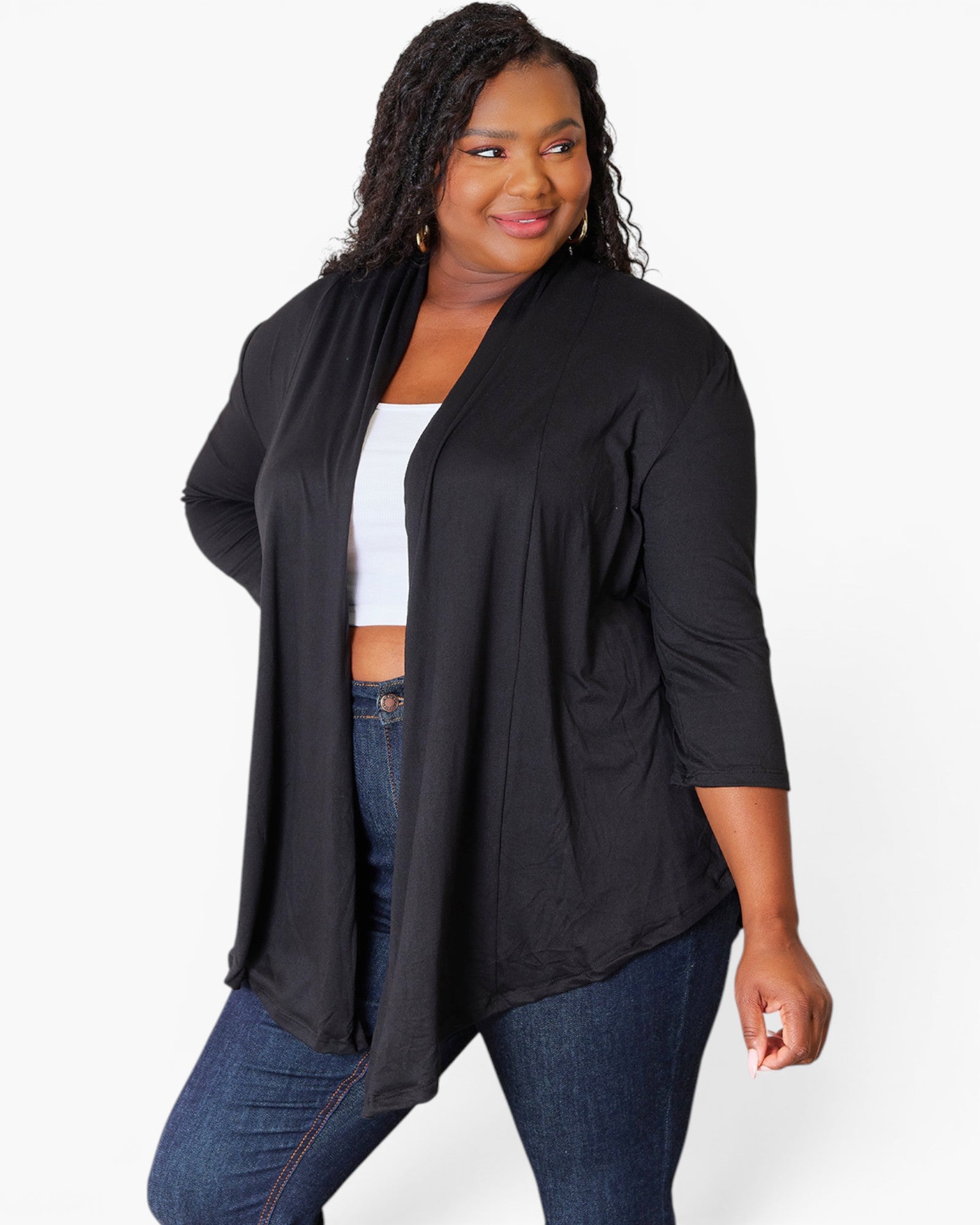 A woman modeling a black open-front cardigan with three-quarter sleeves, worn over a white crop top and blue jeans, against a plain white studio background.