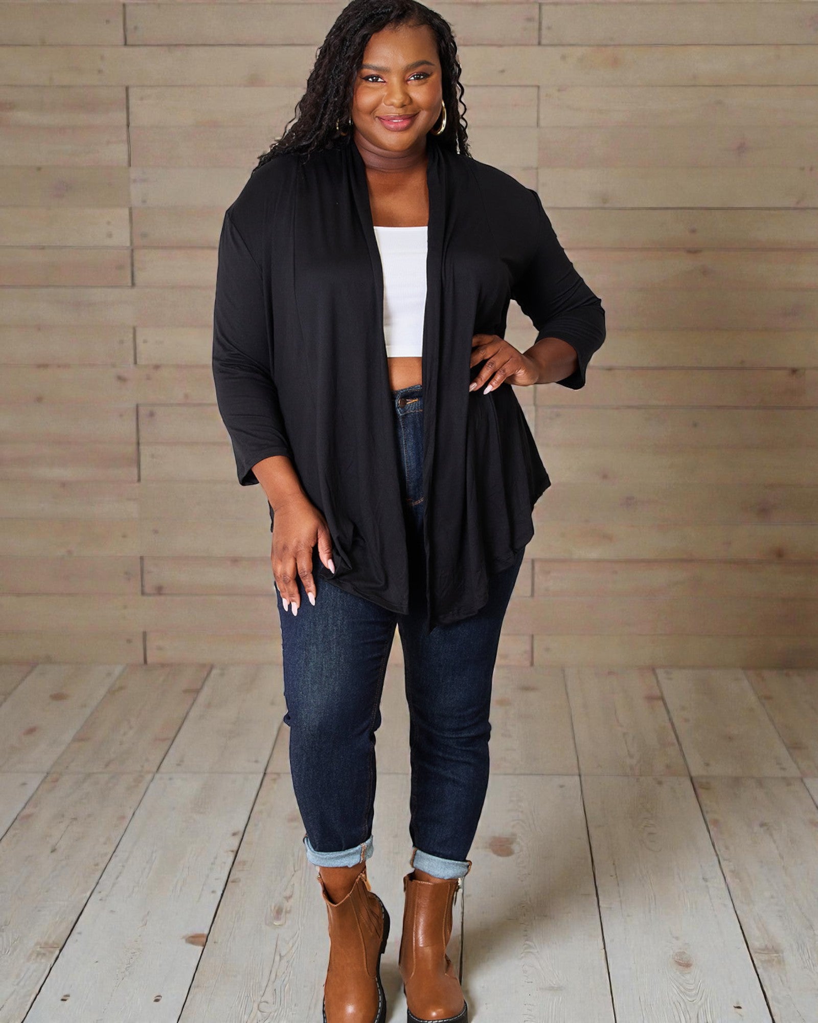 Woman wearing a black cardigan, white top, blue jeans, and brown boots against a wooden background