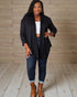 Woman wearing a black cardigan, white top, blue jeans, and brown boots against a wooden background