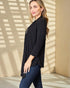 Woman wearing a black top and blue jeans standing against a beige wall with sunlight casting shadows.