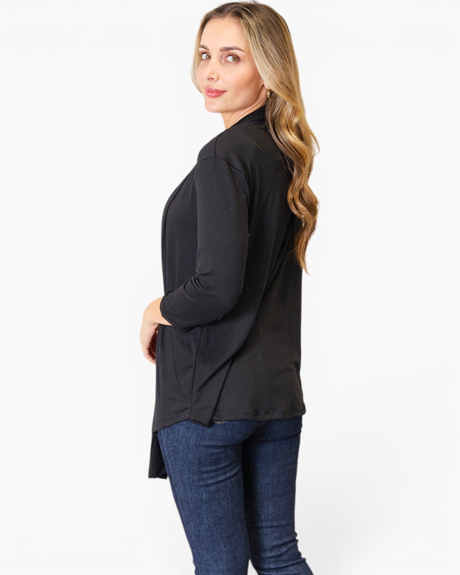 A woman modeling a black long-sleeve wrap-style top with a high-low hem, paired with blue jeans, shown from a side-back angle against a plain white studio background.