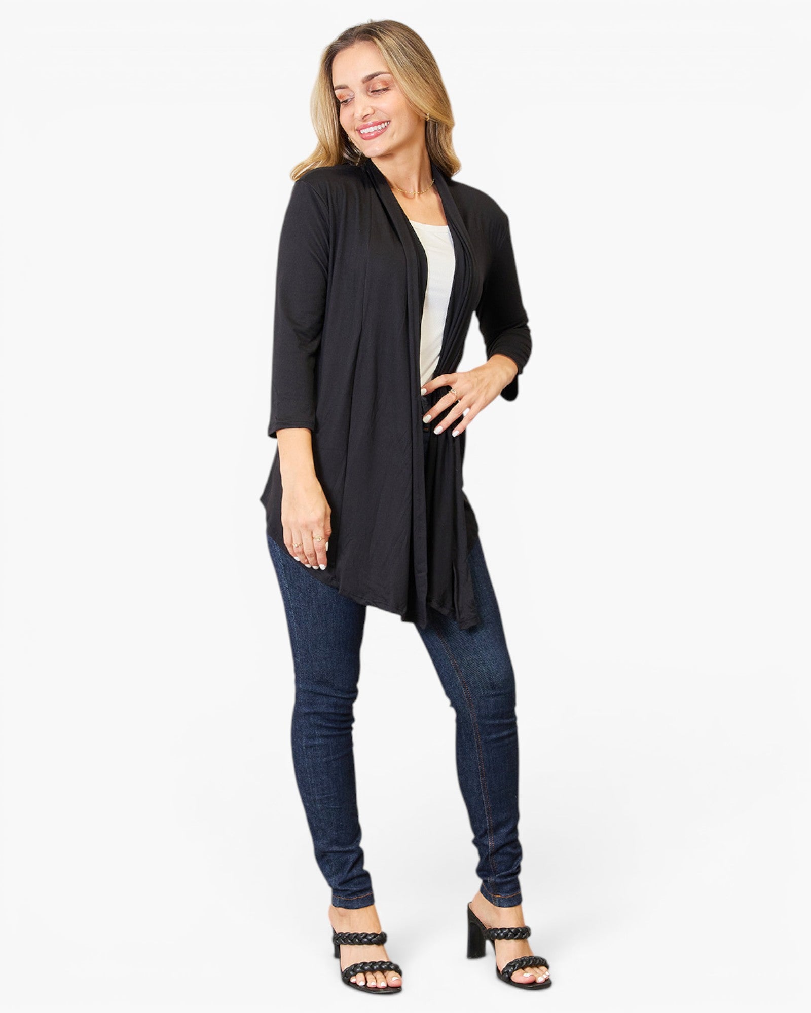 A woman modeling a black open-front longline cardigan over a white top, paired with dark blue skinny jeans and black heeled sandals, against a plain white studio background.