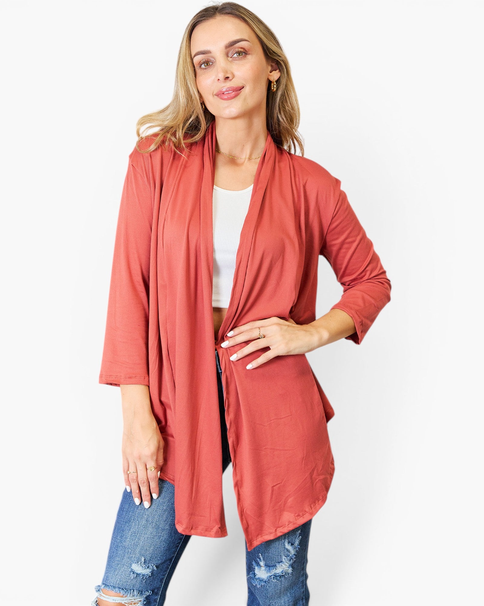 A woman modeling a rust-colored, open-front cardigan with three-quarter sleeves over a white top and blue jeans, set against a plain white studio background.