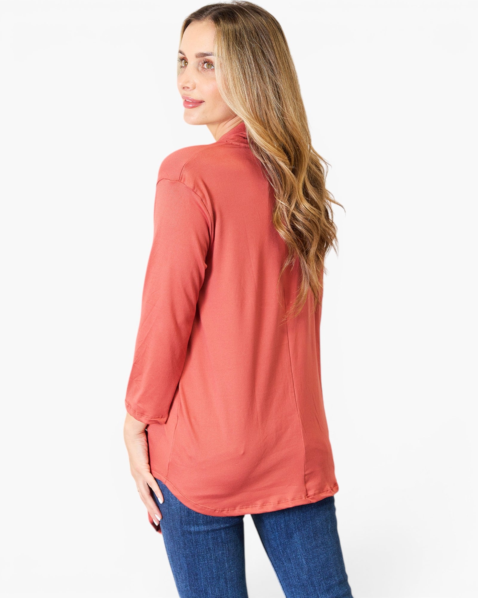 A woman with long blonde hair is shown from a side-back angle wearing a coral pink long-sleeve top and blue jeans against a plain white background.