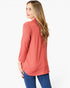 A woman with long blonde hair is shown from a side-back angle wearing a coral pink long-sleeve top and blue jeans against a plain white background.