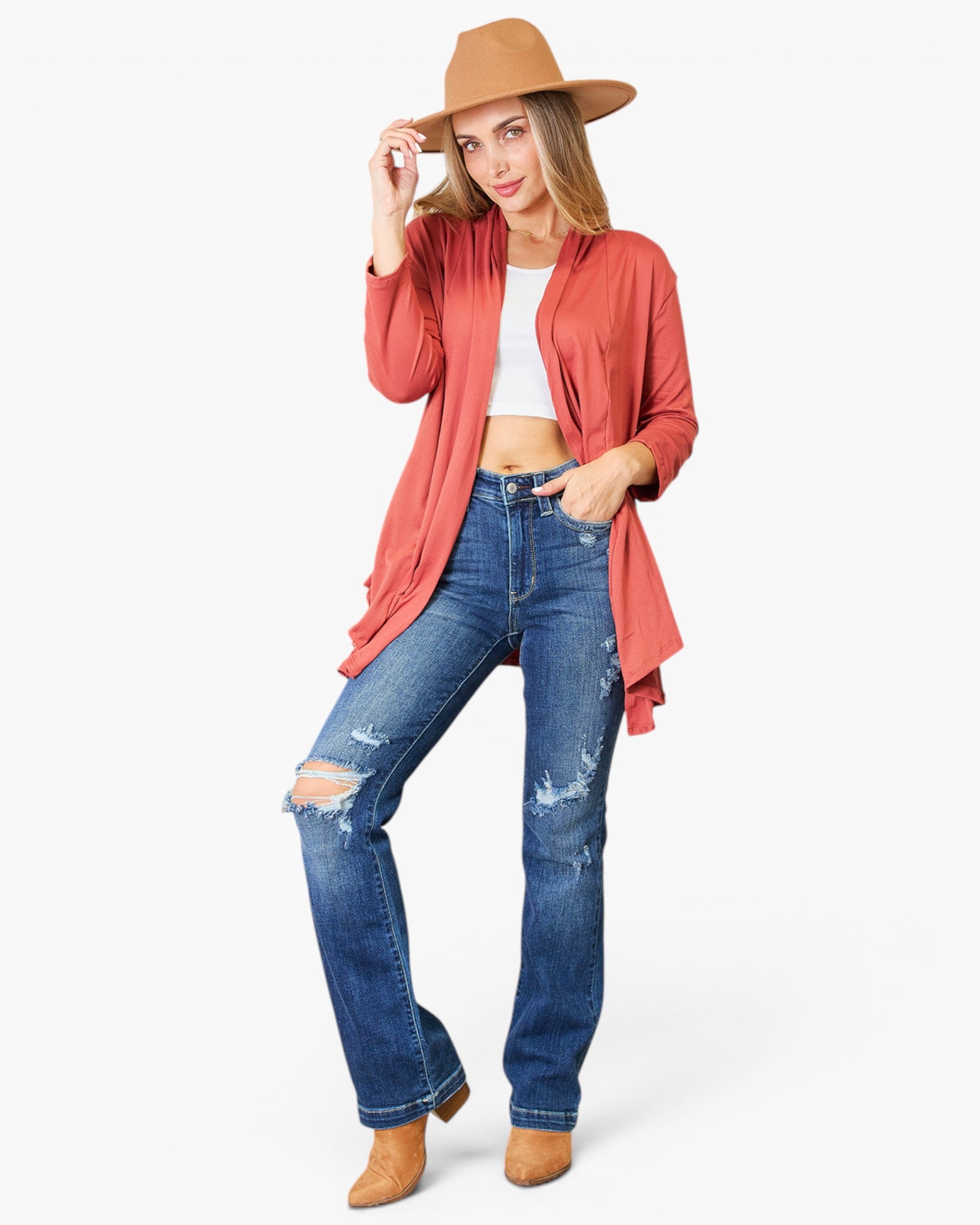 A woman modeling a rust-colored open-front cardigan over a white crop top, paired with distressed blue jeans and tan boots, on a light gray background.