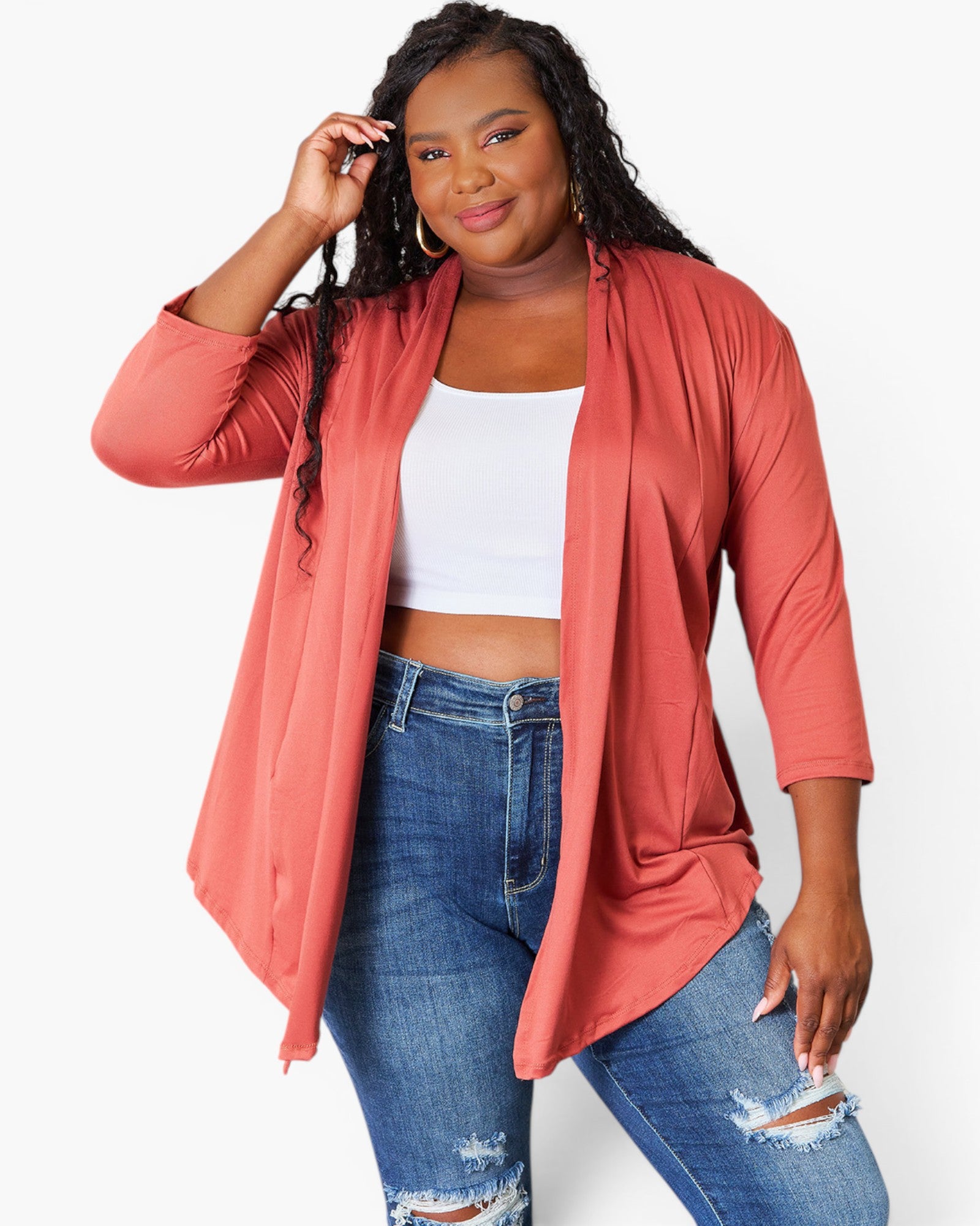 A plus-size model wearing a coral pink open-front cardigan over a white crop top and blue distressed jeans, posed against a light gray studio background.