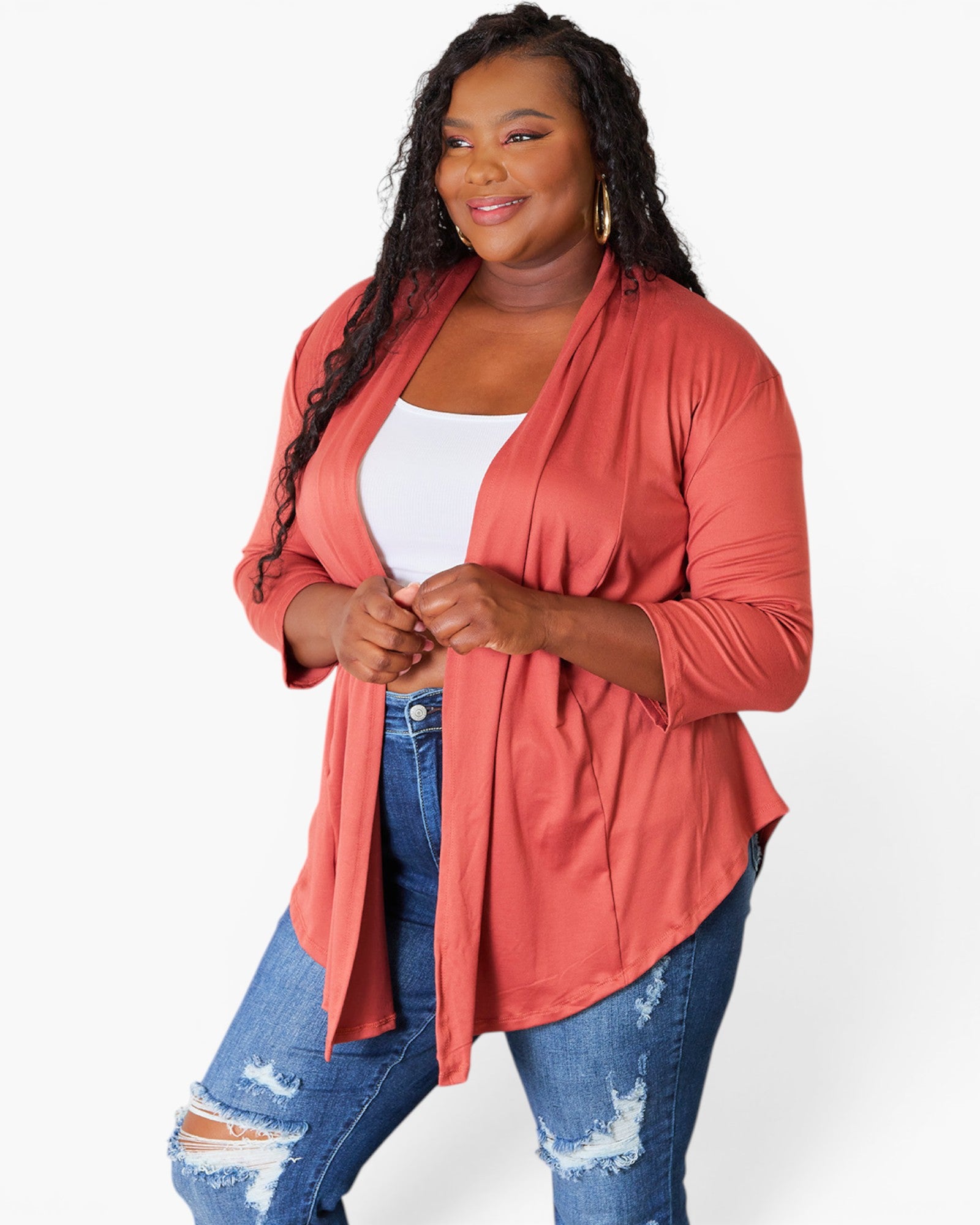 A plus-size woman modeling a coral pink open-front cardigan over a white tank top and distressed blue jeans, photographed against a clean white studio background.
