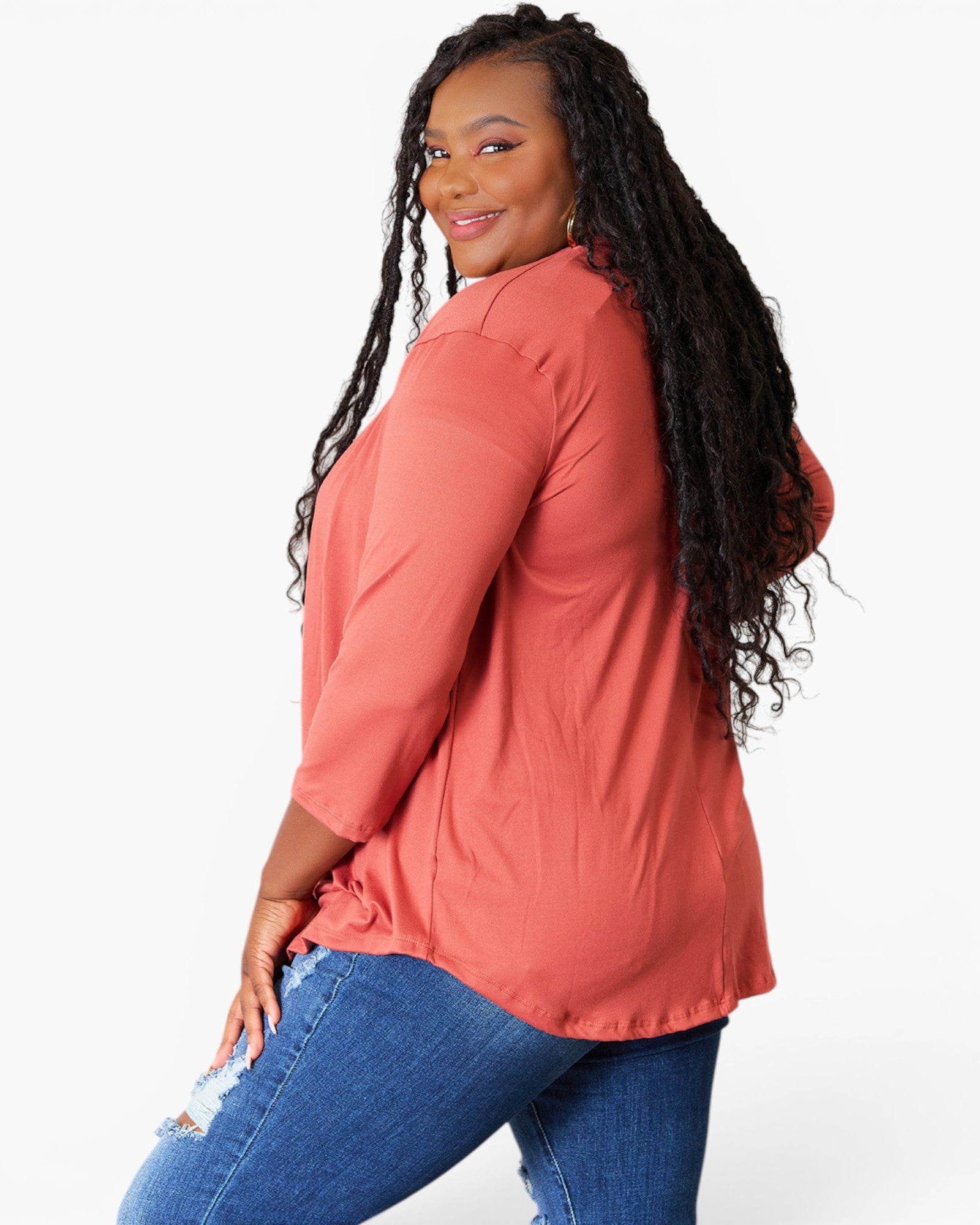 A woman modeling a three-quarter sleeve coral pink button-up shirt with blue jeans, shown from a three-quarters back view against a plain white background.