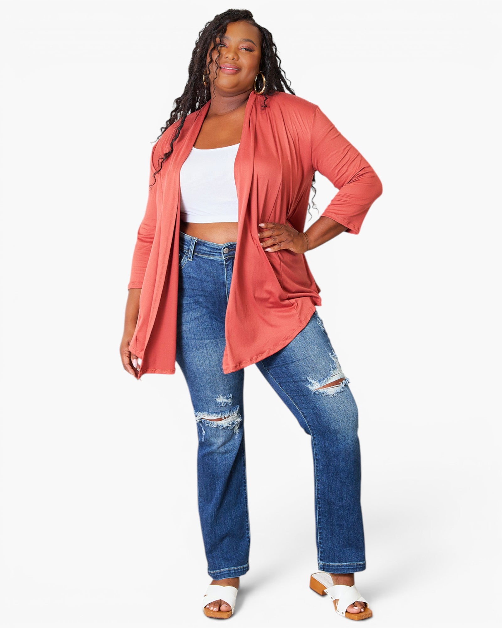 A woman modeling a coral pink open-front cardigan over a white crop top and distressed blue jeans, standing against a light gray studio background.