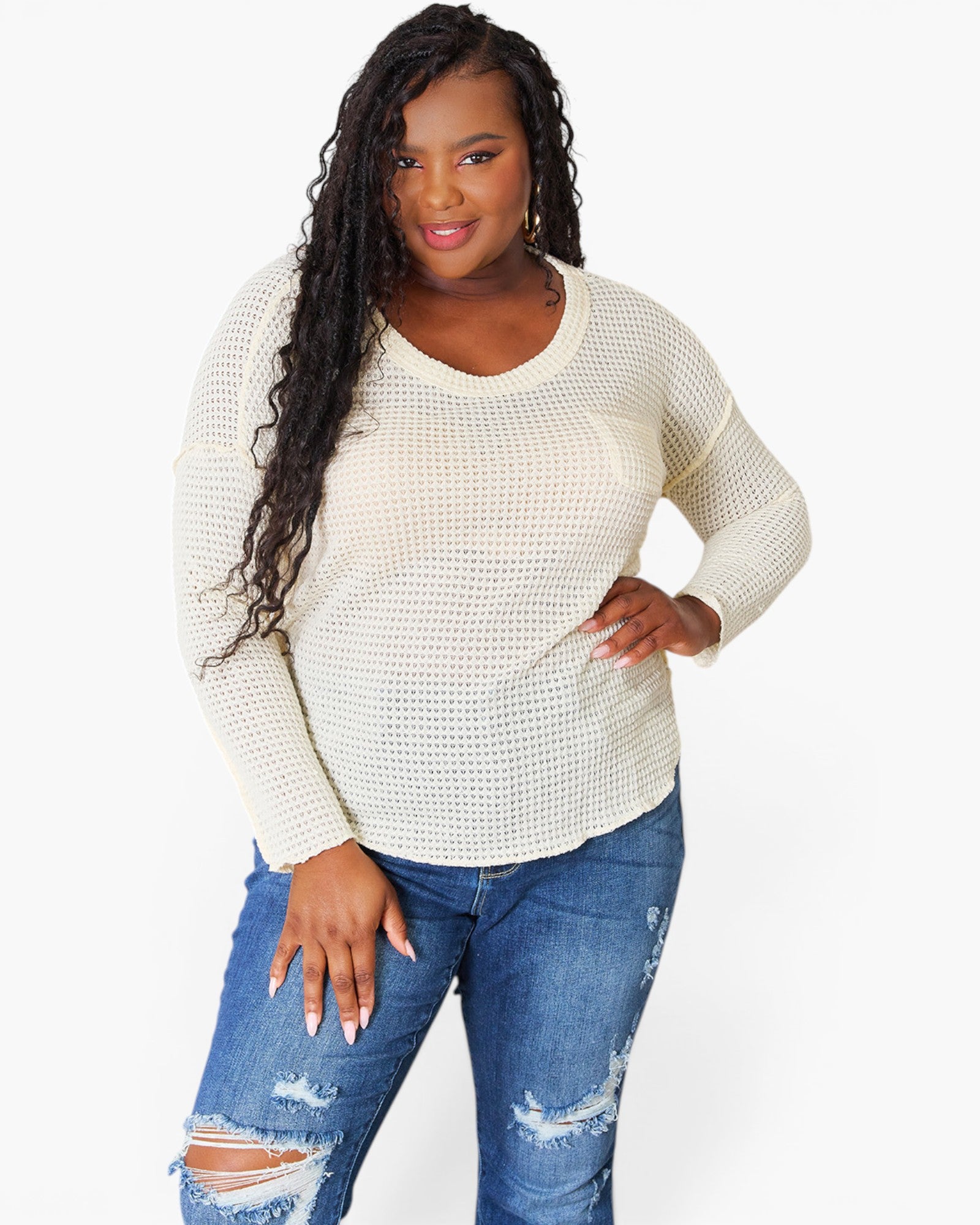 A woman modeling a cream-colored, long-sleeve waffle-knit sweater with a chest pocket, paired with distressed blue jeans, against a plain white background.