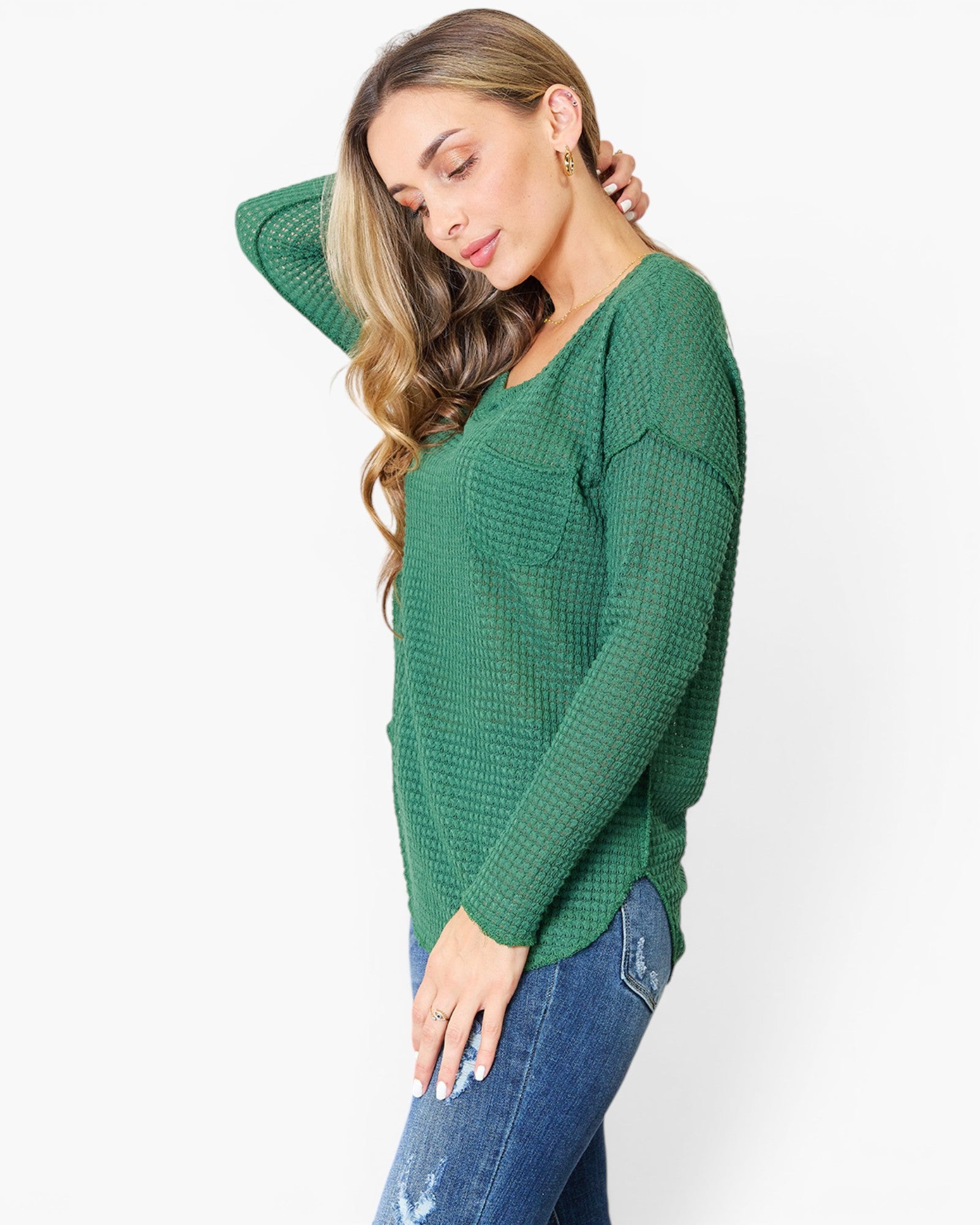 A woman wearing a green waffle-knit long-sleeve top and blue jeans, posing against a plain white studio background.