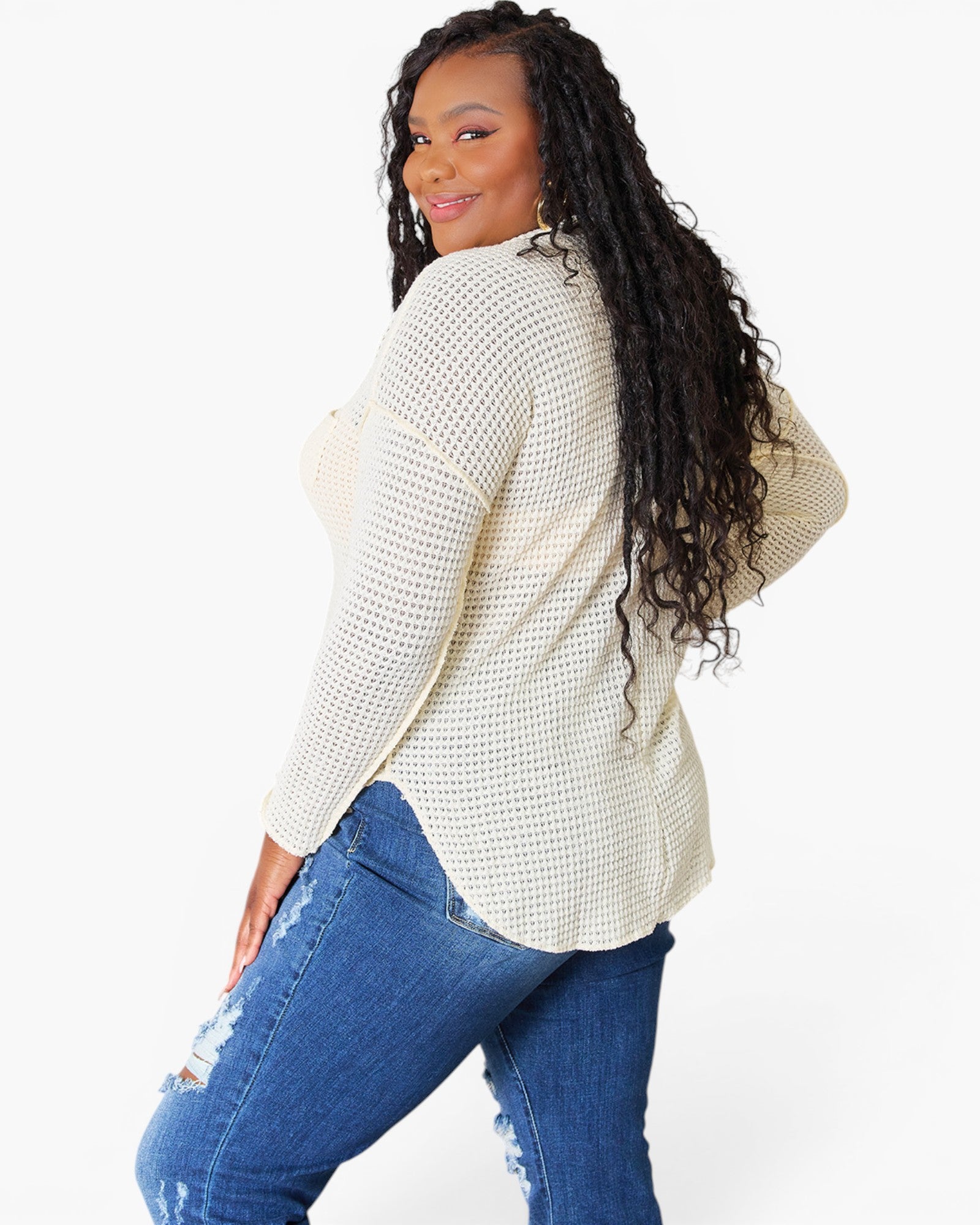 A woman modeling a cream-colored, long-sleeve open-knit top with blue distressed jeans against a plain white studio background.