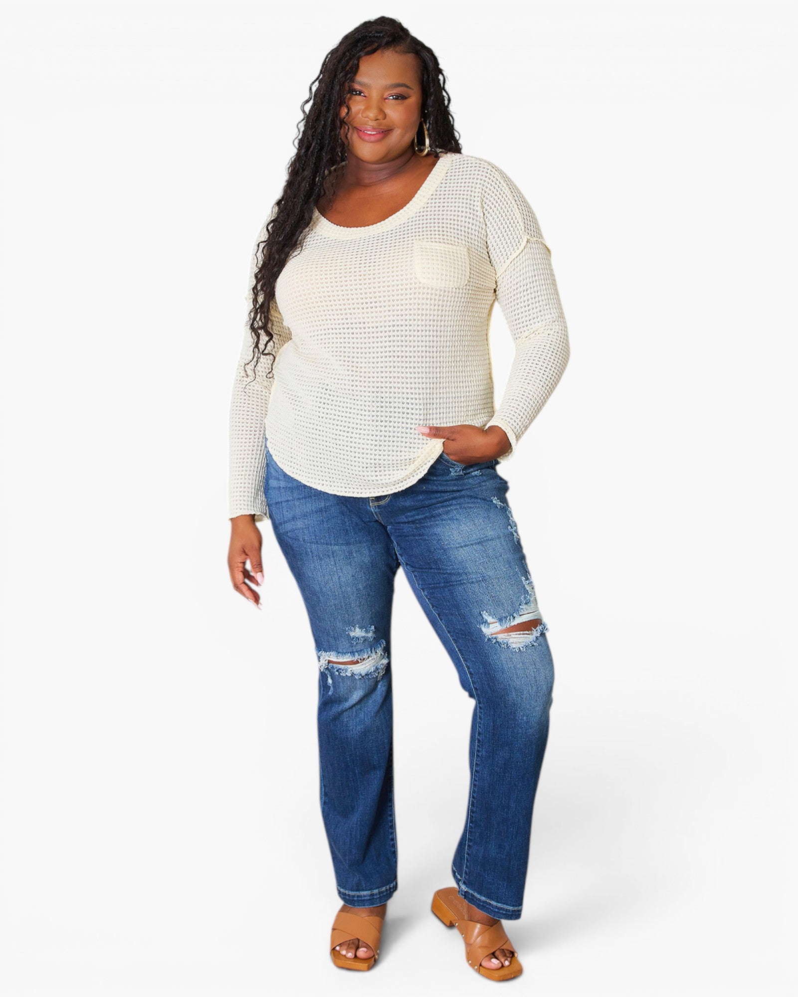 A woman modeling a cream-colored, textured long-sleeve top with a chest pocket, paired with distressed blue jeans and tan sandals, against a plain white studio background.