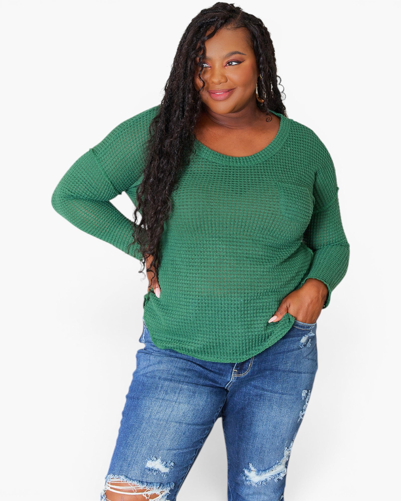 A woman modeling a green waffle-knit long-sleeve top with blue distressed jeans against a plain white background.