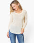 A woman wearing a cream-colored, long-sleeve waffle-knit top with a chest pocket and blue jeans, photographed against a plain white studio background.