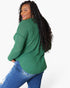 A woman with long black curly hair is shown from a side-back angle wearing a green waffle-knit long-sleeve top and blue jeans against a plain white background.