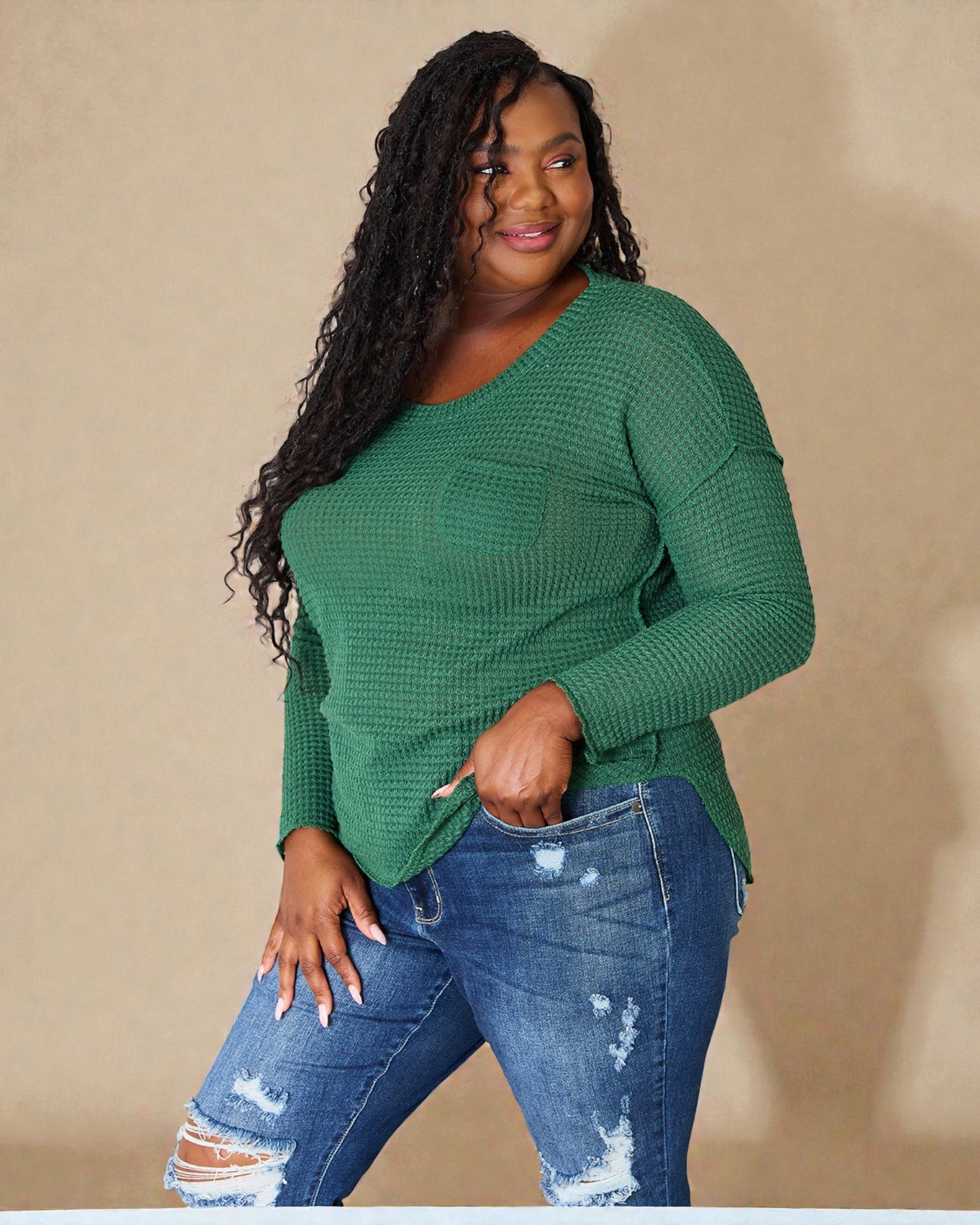 Woman wearing a green long-sleeve top and blue jeans against a beige background