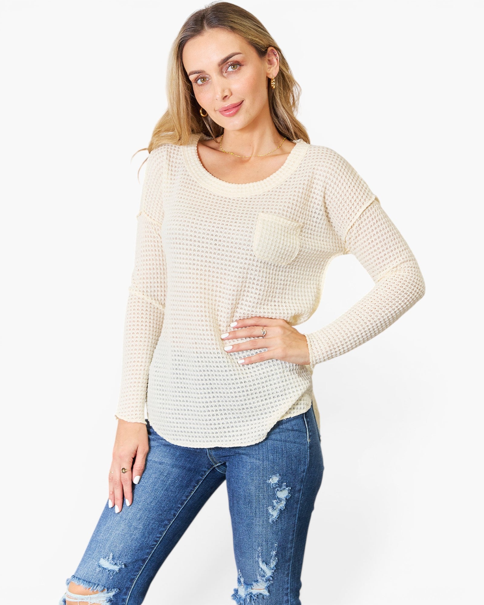 A woman modeling a cream-colored, long-sleeve waffle-knit top with a chest pocket, paired with distressed blue jeans, against a plain white studio background.