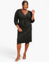 A woman modeling a black ribbed wrap-style midi dress with three-quarter sleeves and a tie waist, photographed against a clean light gray studio background.