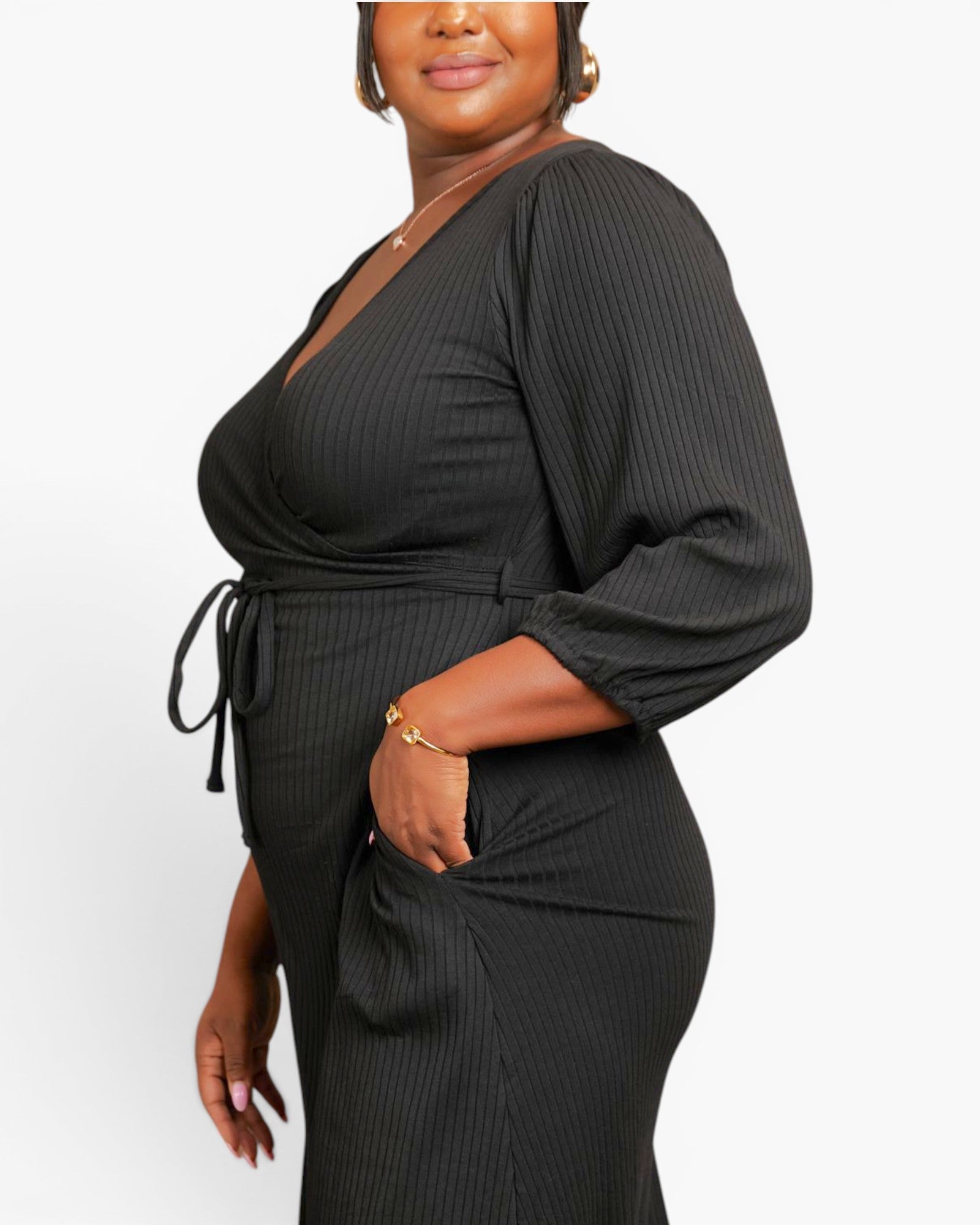 A plus-size woman wearing a black ribbed wrap-style jumpsuit with three-quarter sleeves and a tie waist, shown from the chest down against a plain light background.