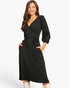 A woman modeling a black ribbed midi wrap dress with three-quarter puff sleeves, side pockets, and a self-tie waist belt, paired with white boots against a plain white studio background.