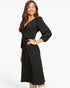 A woman modeling a black ribbed midi wrap dress with three-quarter sleeves and a self-tie waist, photographed against a clean white studio background.