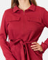 A close-up of a woman wearing a long-sleeve collared knit top or lightweight dress in a rich red color, featuring a half-zip neckline, two chest pockets, and a matching waist tie.