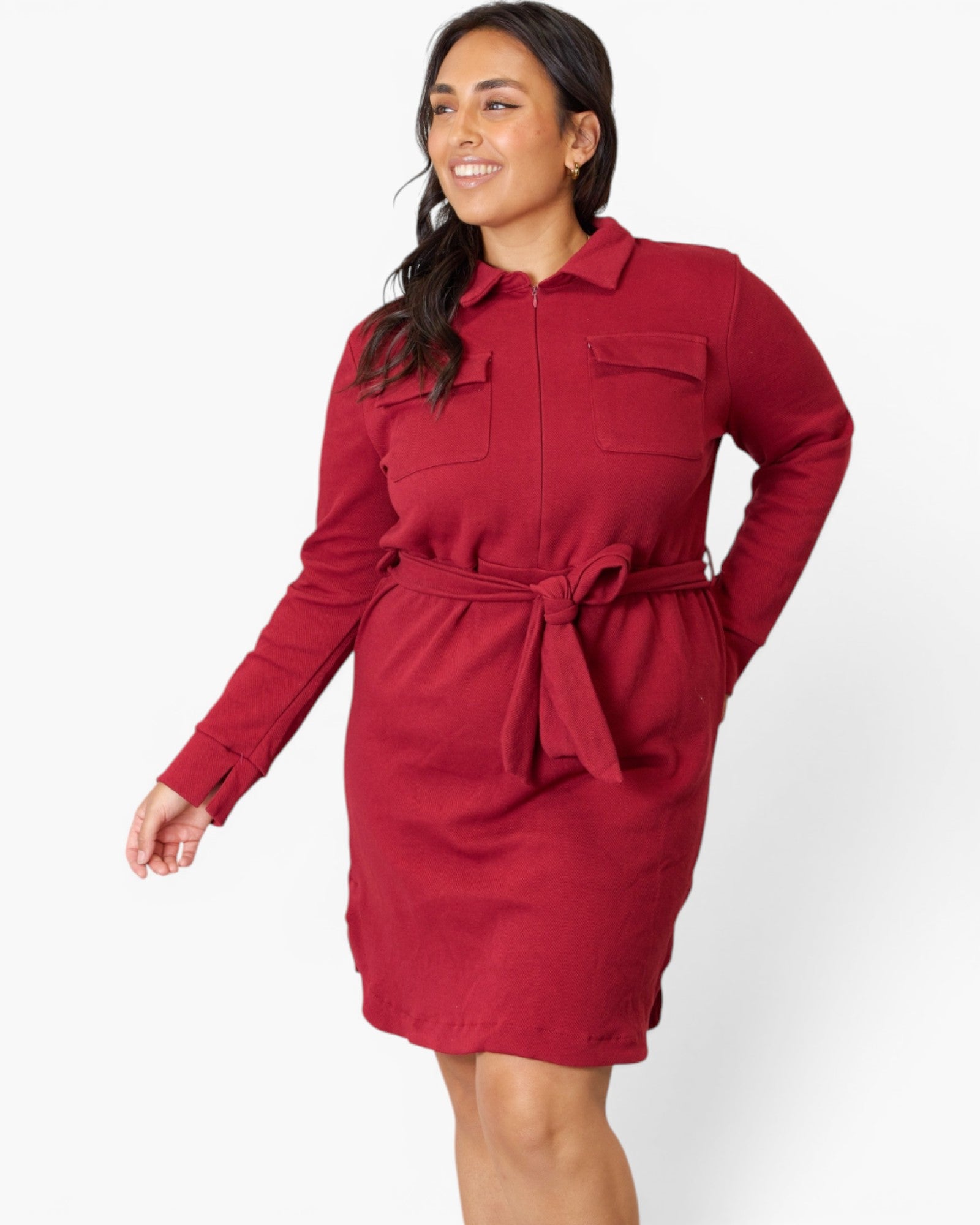 A woman wearing a long-sleeve red collared shirt dress with a matching tie belt, shown against a plain light gray studio background.