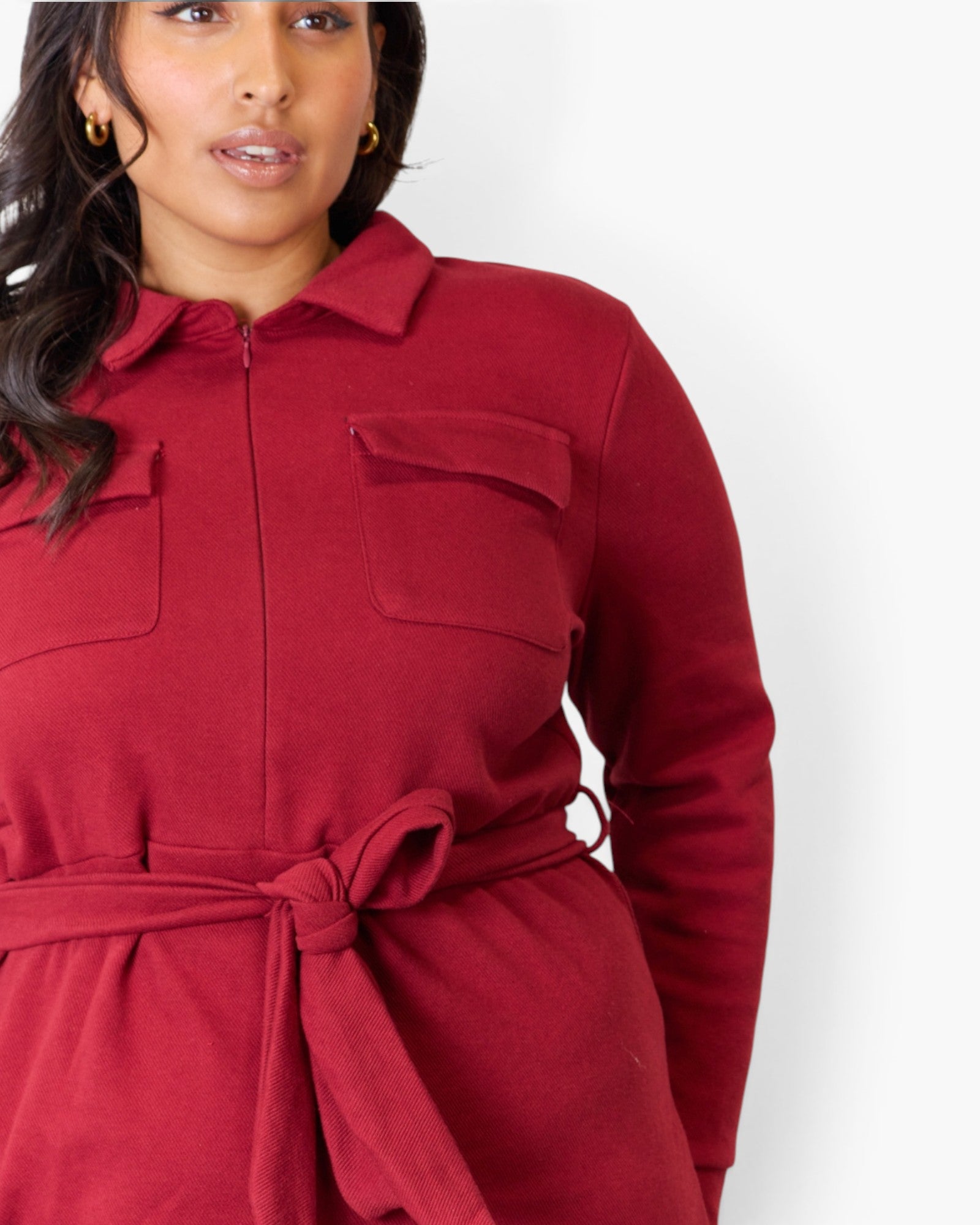 A woman wearing a long-sleeve collared jumpsuit in deep burgundy red, featuring a zip-up front, chest pockets, and a matching tie belt at the waist.