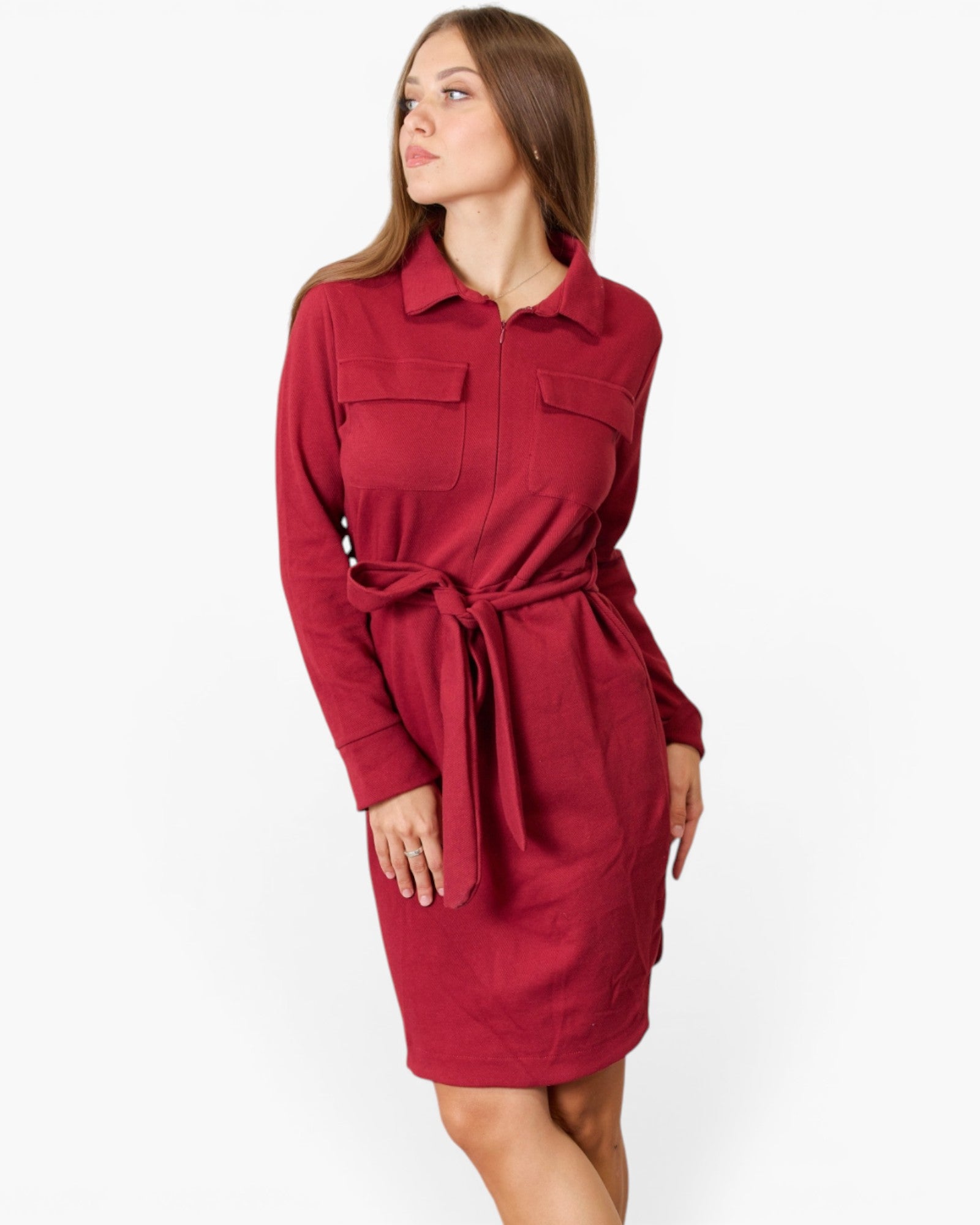 A woman modeling a long-sleeve collared shirt dress in deep red, featuring a matching tie belt at the waist and flap chest pockets, set against a plain light gray studio background.