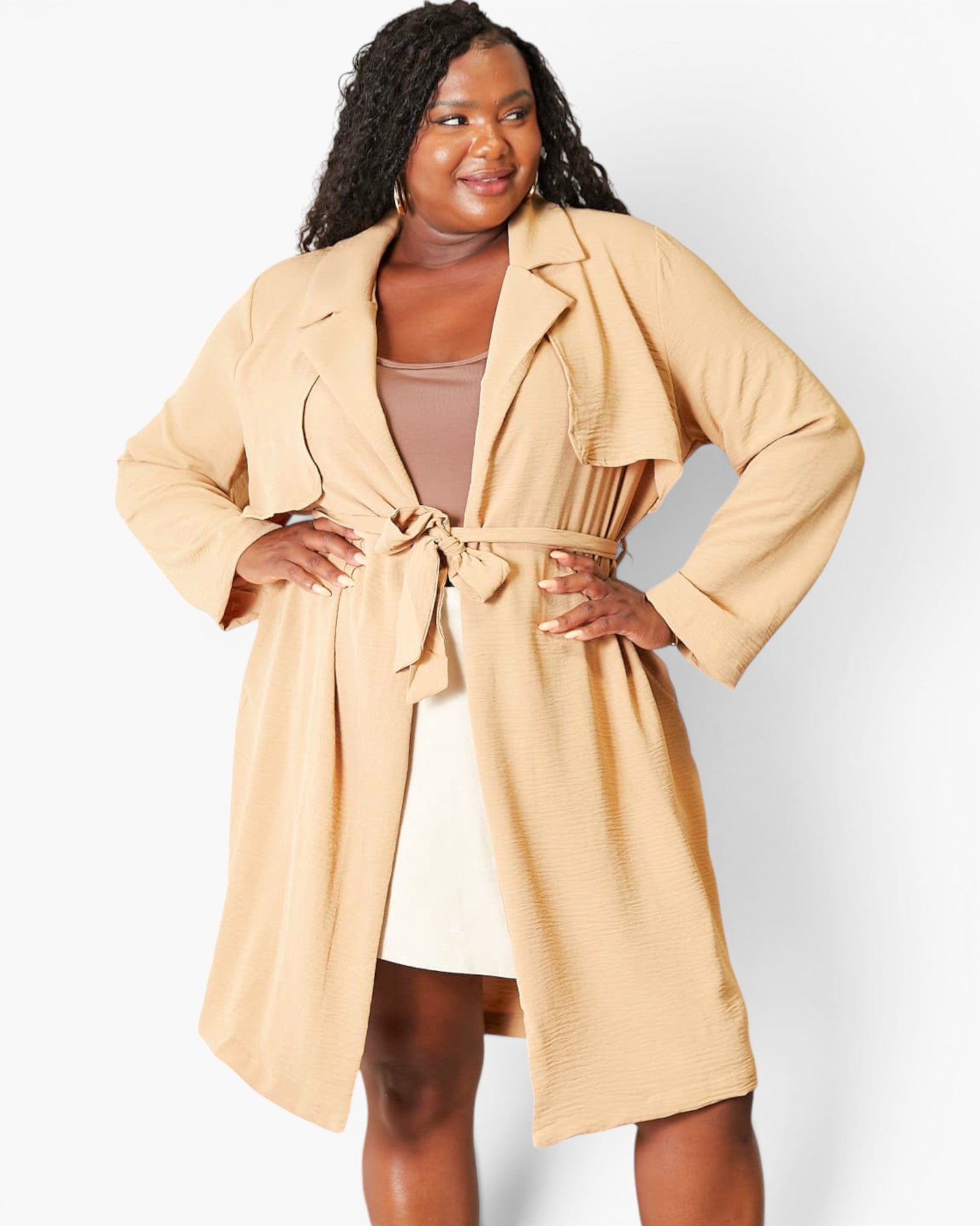 A woman modeling a light beige waffle-knit longline wrap coat with a tie belt, worn over a beige top and cream skirt against a light gray studio background.