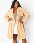 A woman modeling a light beige waffle-knit longline wrap coat with a tie belt, worn over a beige top and cream skirt against a light gray studio background.