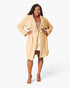 A woman modeling a light tan belted trench coat over a beige dress, standing against a plain white studio background.