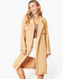 A woman modeling a long, light tan trench coat over a brown top and white skirt, photographed against a clean white studio background.