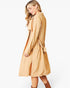 A woman wearing a light tan trench coat with a matching waist tie belt, shown from a side-back angle against a plain white studio background.