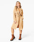 A woman modeling a tan trench coat with a matching tie belt, worn over a brown top and paired with brown ankle boots, against a plain white studio background.