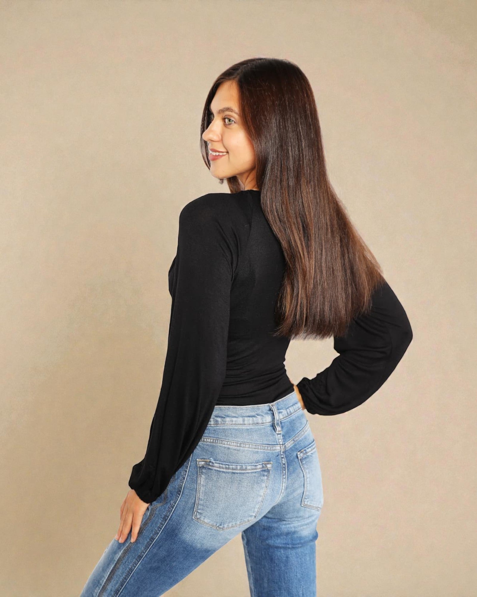 Woman wearing a black long-sleeve top and blue jeans against a beige background
