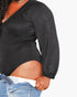 A close-up of a person wearing a black long-sleeve bodysuit or top with gathered sleeves, paired with blue jeans against a plain light background.