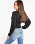 A woman with long brown hair is shown from a side-back angle wearing a fitted black long-sleeve mock-neck top and light blue jeans against a plain white studio background.