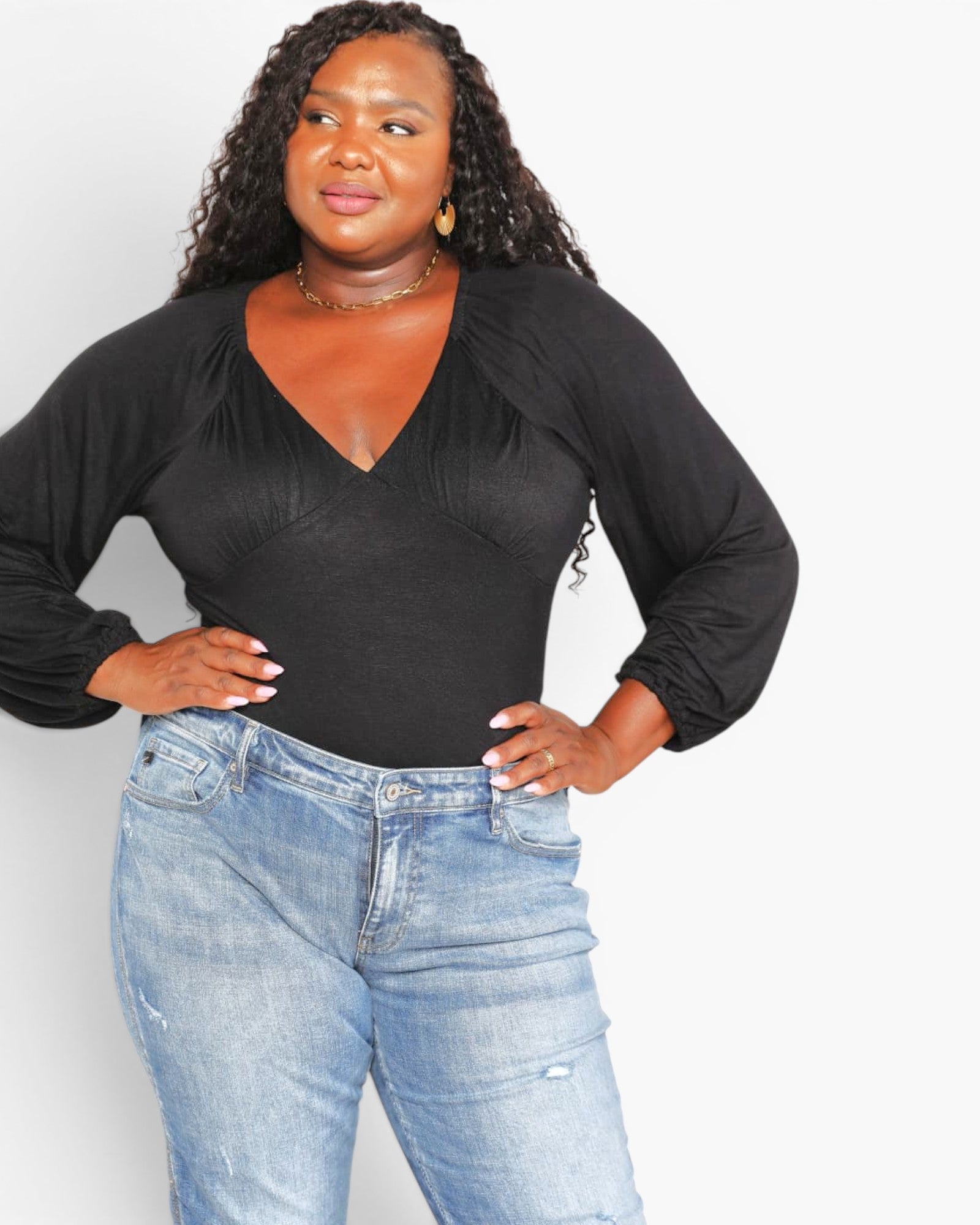 A woman modeling a black long-sleeve wrap-style top with gathered puff sleeves and a V-neckline, paired with light-wash blue jeans against a plain light gray background.