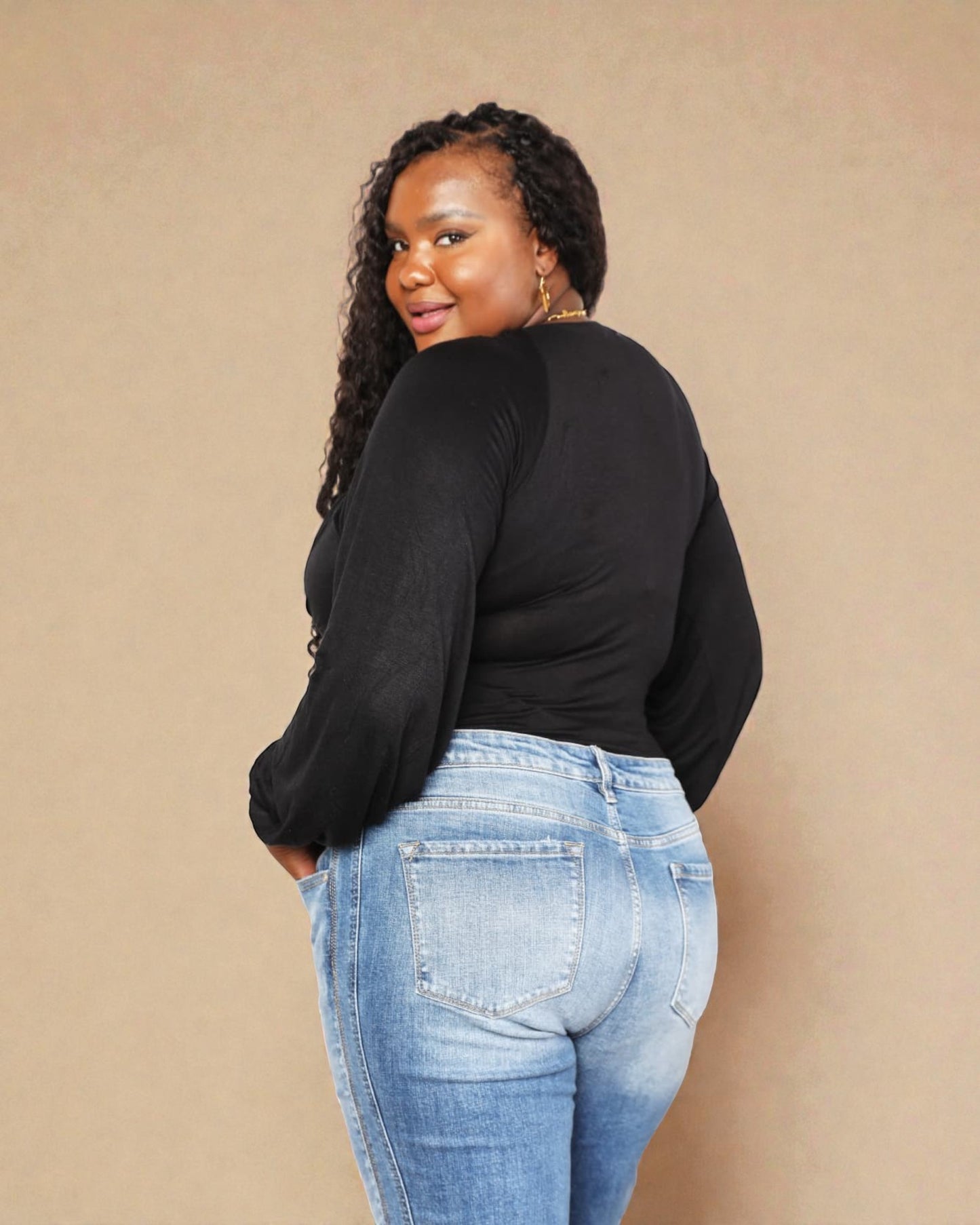Woman wearing a black long-sleeve top and blue jeans against a beige background