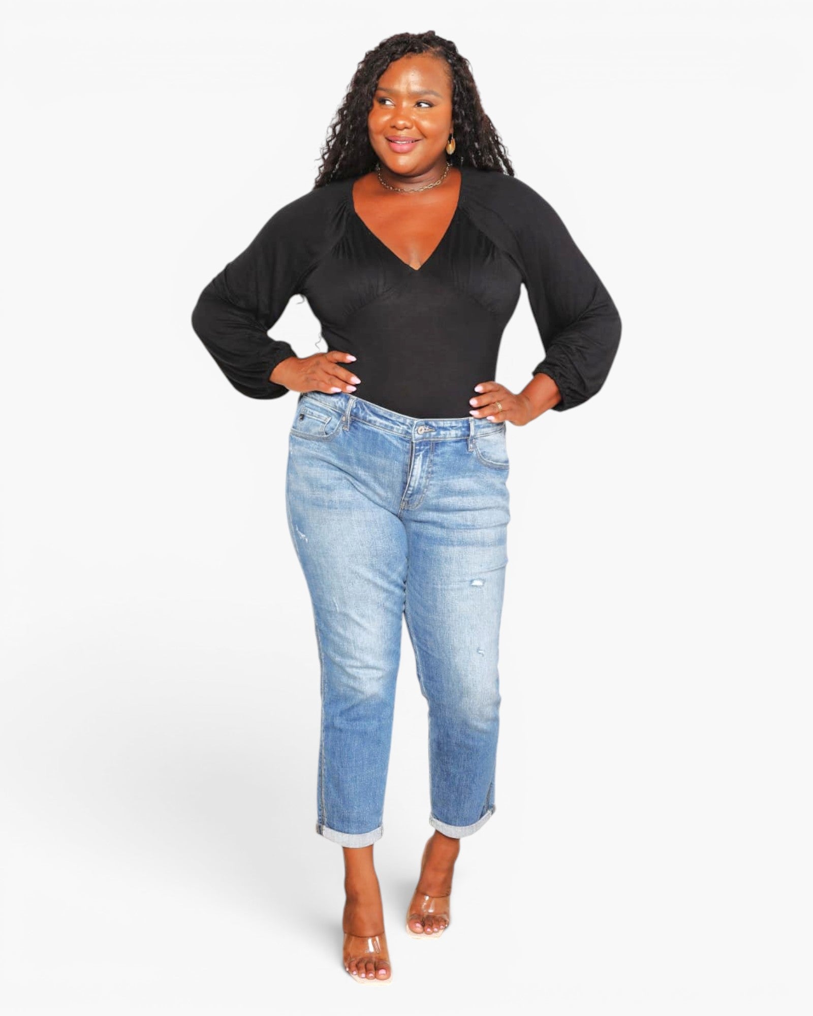 A woman modeling a black long-sleeve V-neck top and light-wash blue cropped jeans with rolled cuffs, standing against a plain light gray studio background.