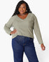 A woman modeling a sage green long-sleeve V-neck top with a front kangaroo pocket, paired with dark blue jeans, against a plain white background.